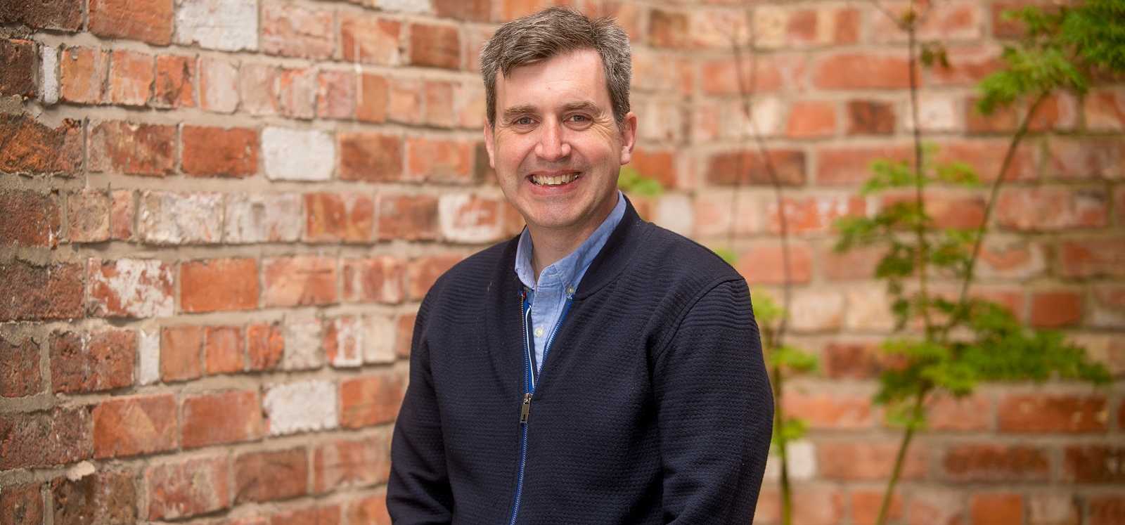 smiling man with thick greying hair, wearing a blue shirt and a navy zip-up jacket, with a red brick wall in the background